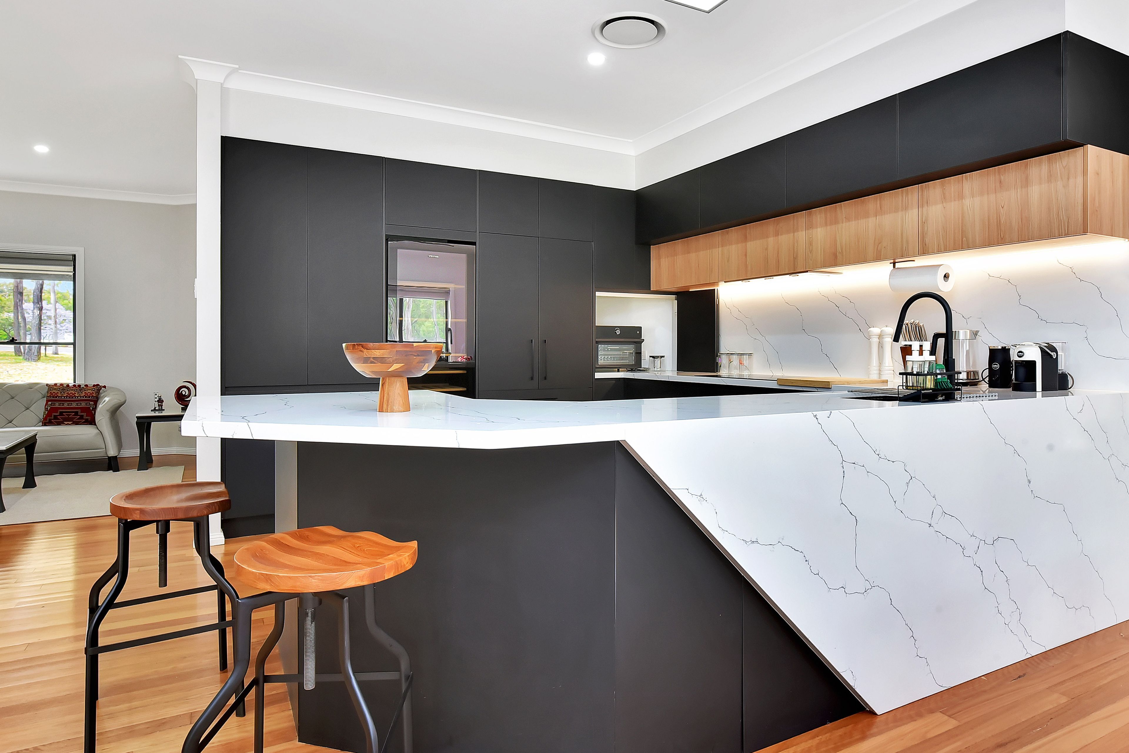 A modern kitchen with a large island, dark cabinetry, and a view to the living area.