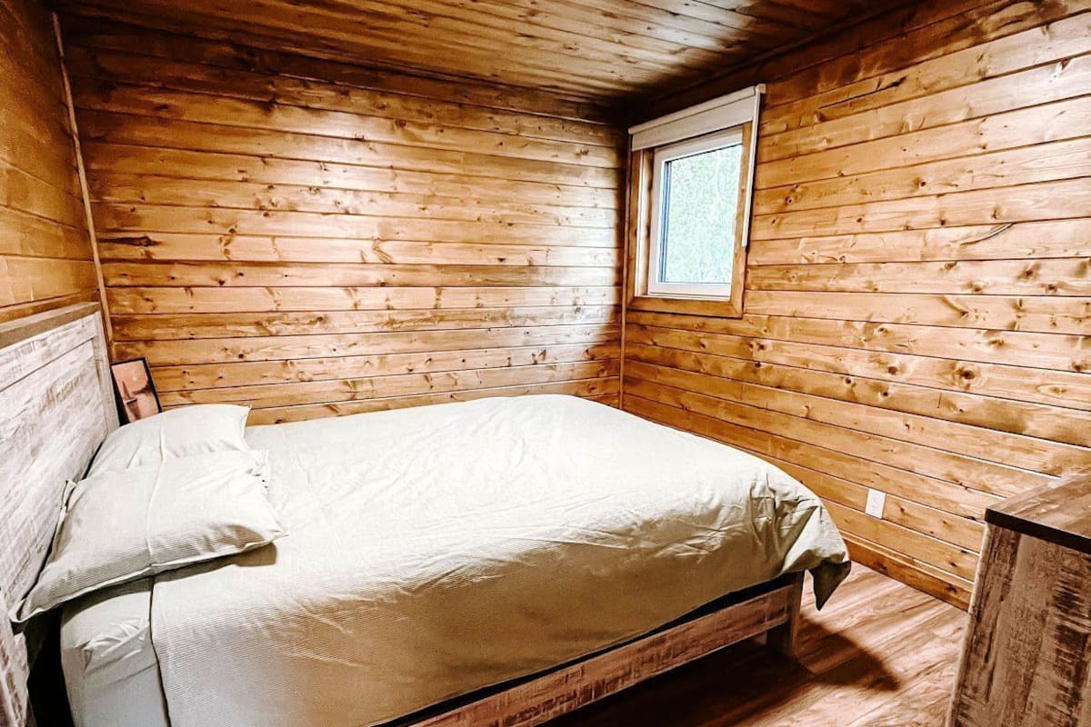 A small, cozy room with a wooden ceiling and walls, a single bed with a mattress and pillows, and a small window.
