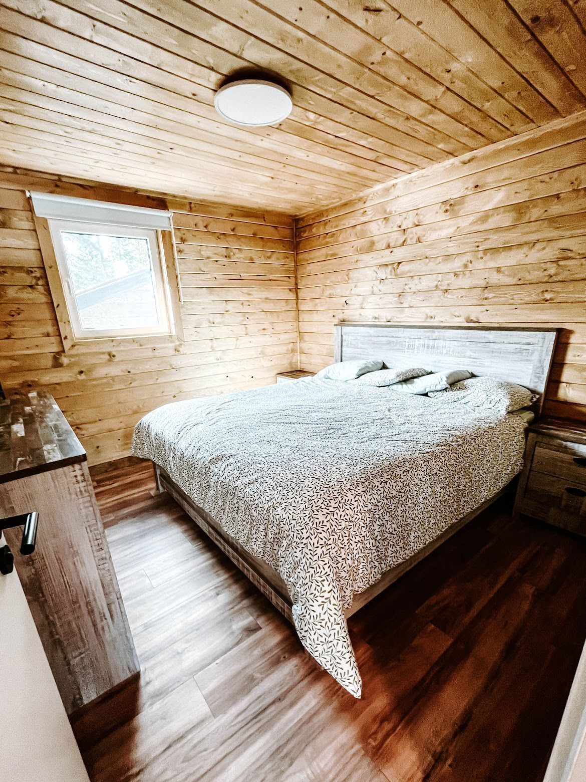 A wooden cabin bedroom with a bed, a nightstand, a window, and a wooden floor.