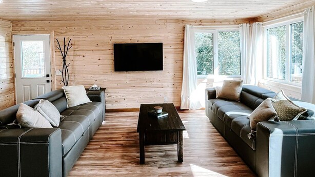 A spacious living room with wooden floors and walls, featuring two dark sofas, a central coffee table, and a large flat-screen TV mounted on the wall. - Faloma Lakeside Cabins (Faloma)