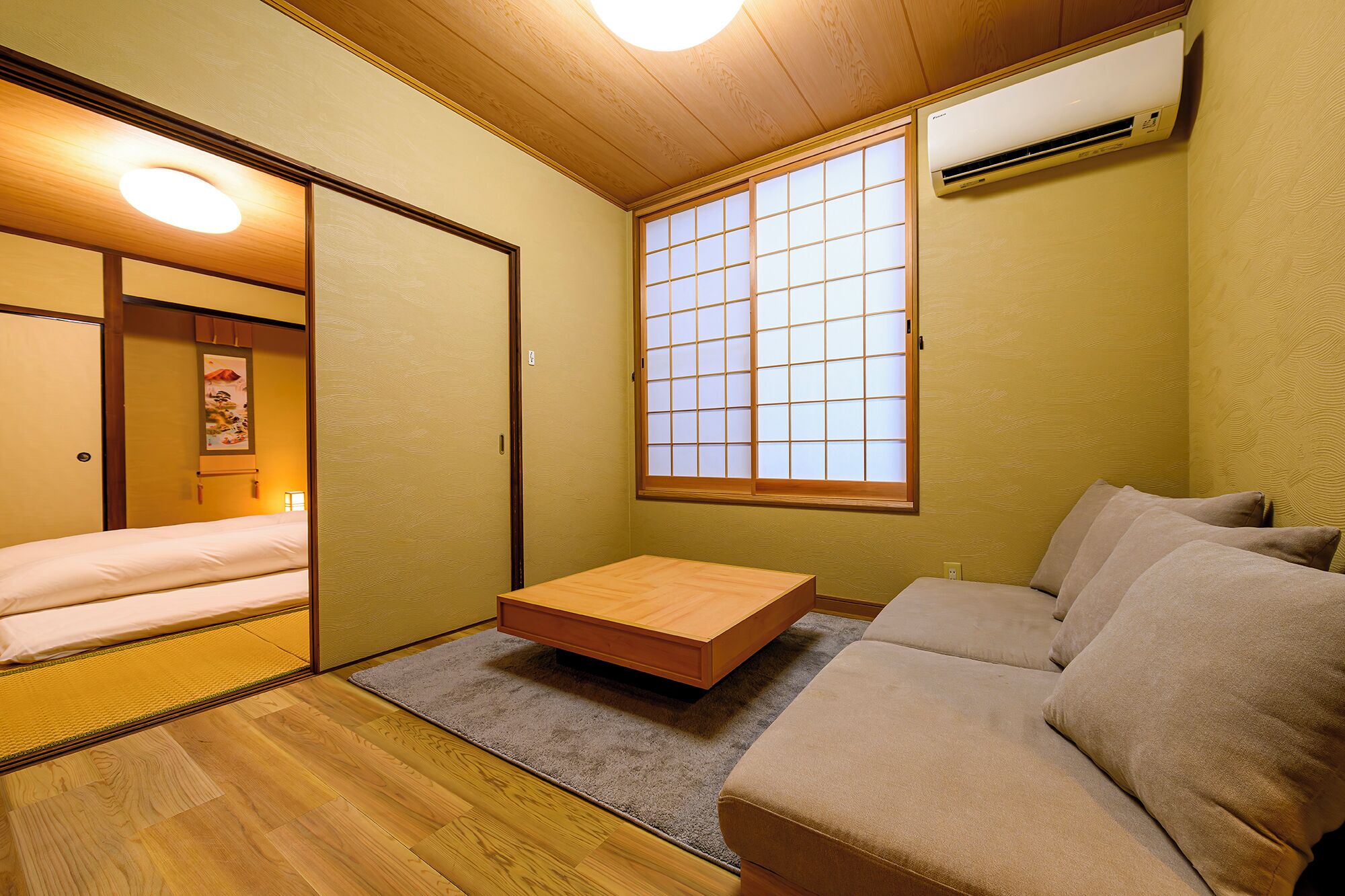 A compact room with a futon bed, a low wooden table, a sofa, a shoji screen window, and an air conditioning unit.