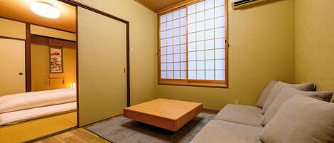 A compact room with a futon bed, a low wooden table, a sofa, a shoji screen window, and an air conditioning unit.