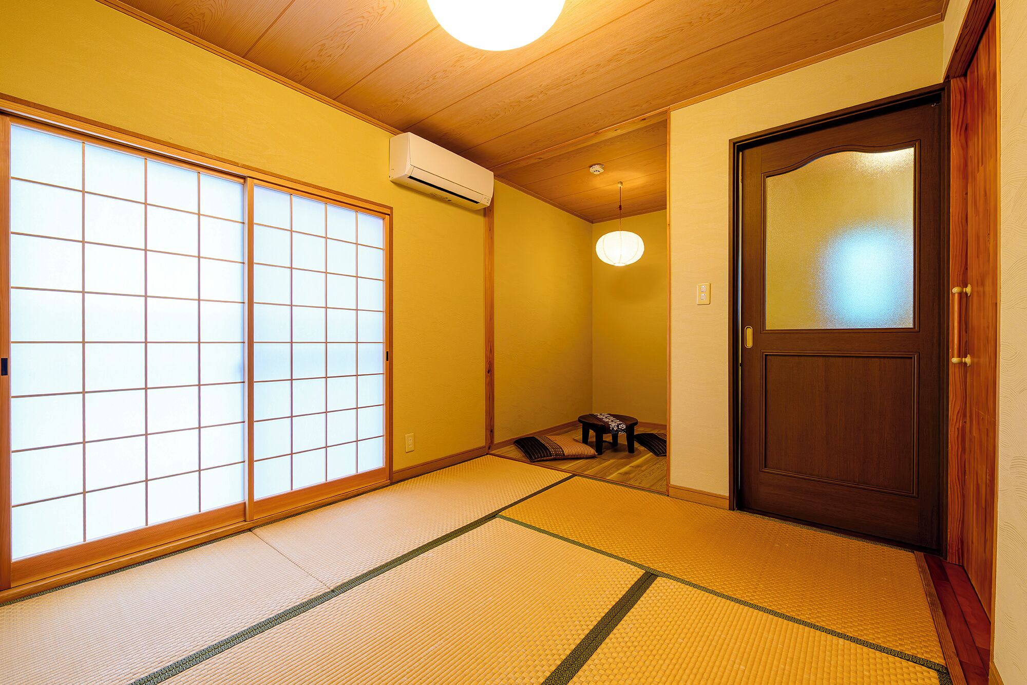 A traditional Japanese room with tatami flooring, a sliding shoji door, a wooden ceiling, and a dark wooden door with a frosted glass panel.