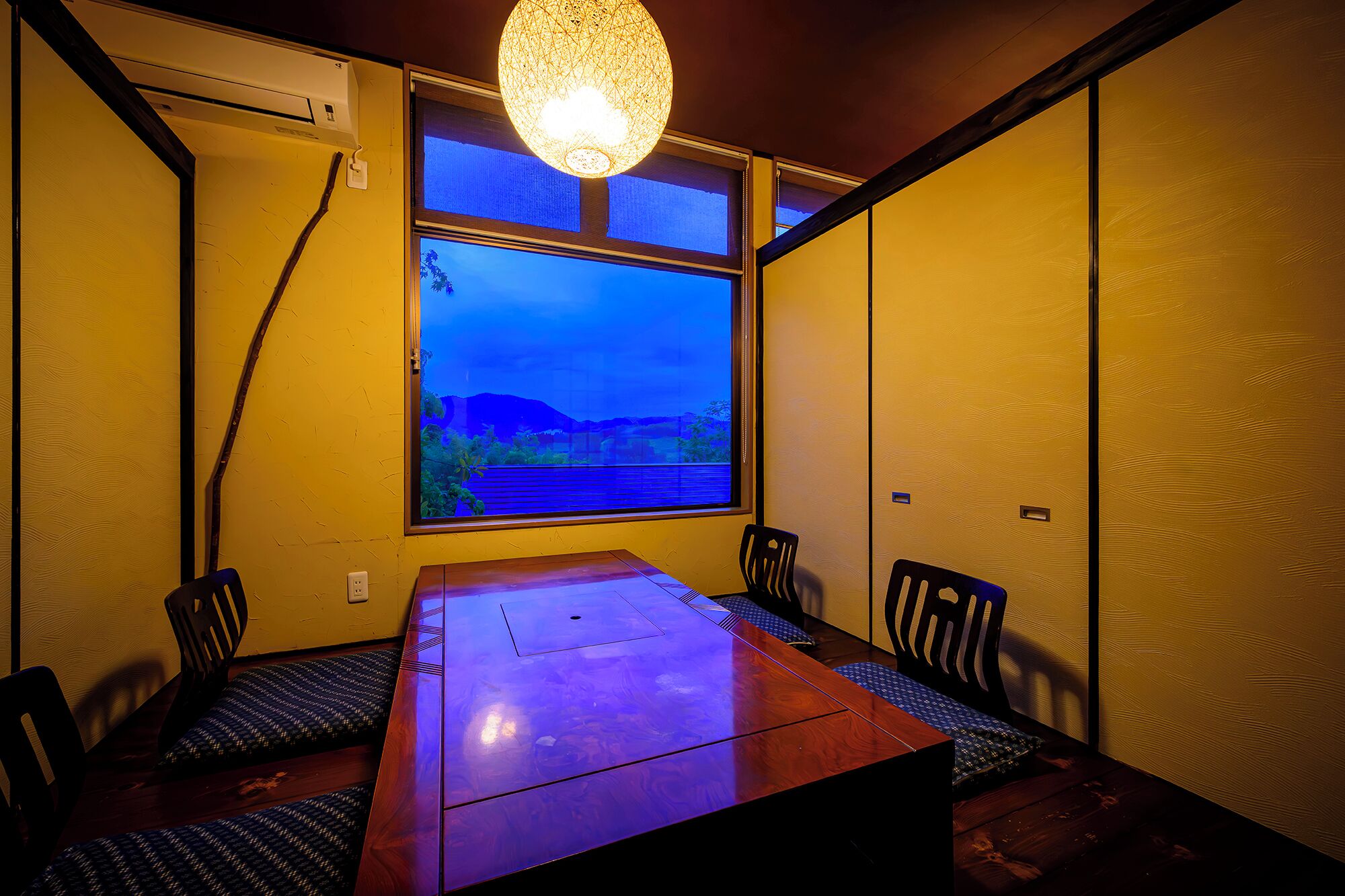 A dining area with a wooden table and chairs, a large window with a view of a landscape, and a hanging light fixture.