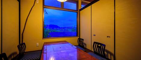 A dining area with a wooden table and chairs, a large window with a view of a landscape, and a hanging light fixture.