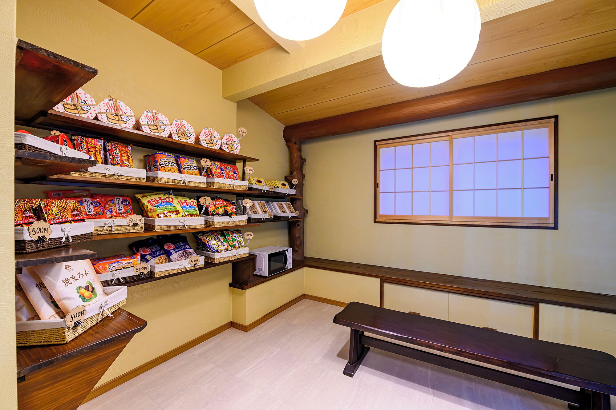 A room with wooden shelves displaying snacks, a bench, a microwave, and a window with a view.