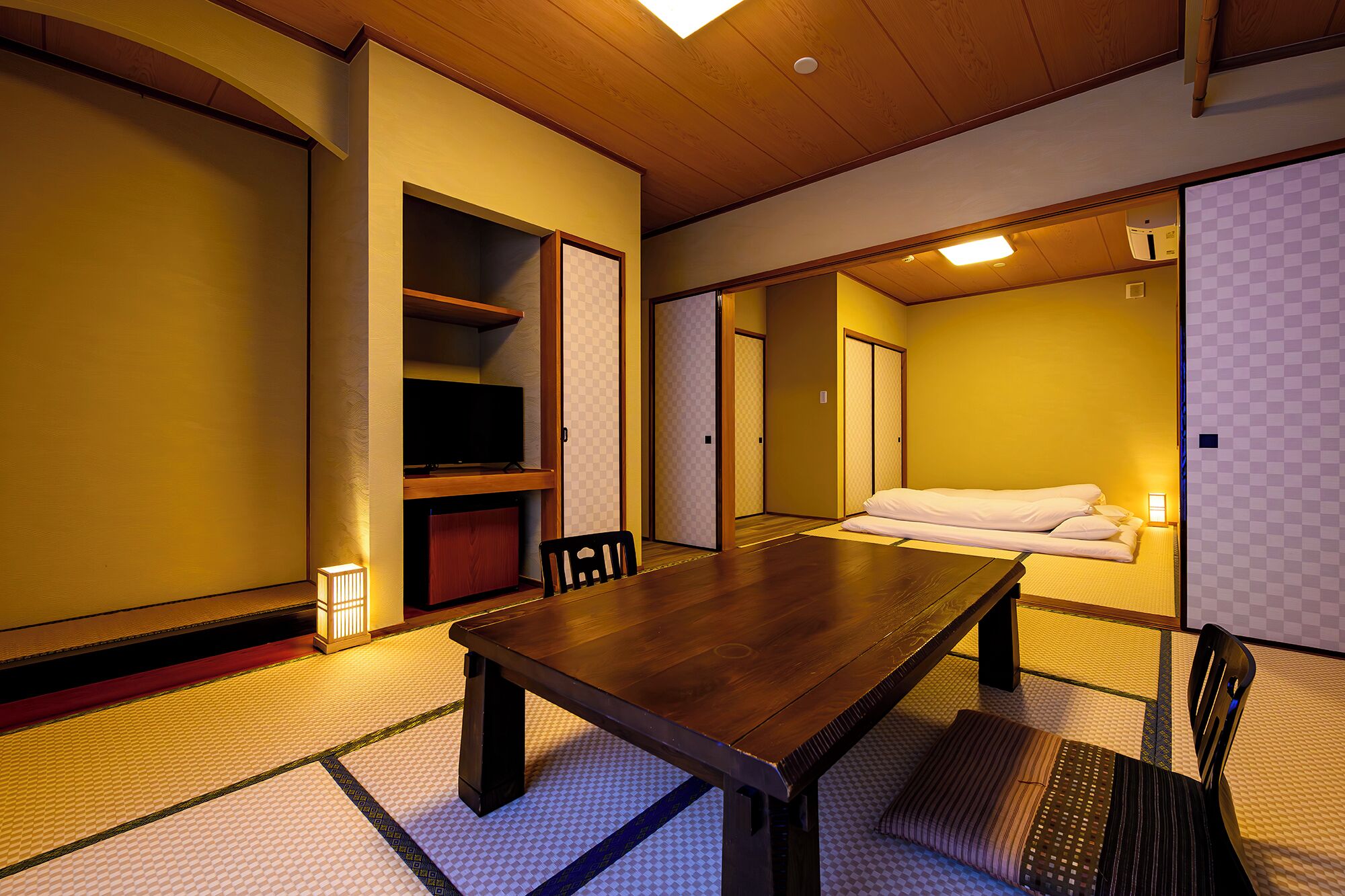 A traditional Japanese room with tatami flooring, a low wooden table, a television, and a bed with white bedding.