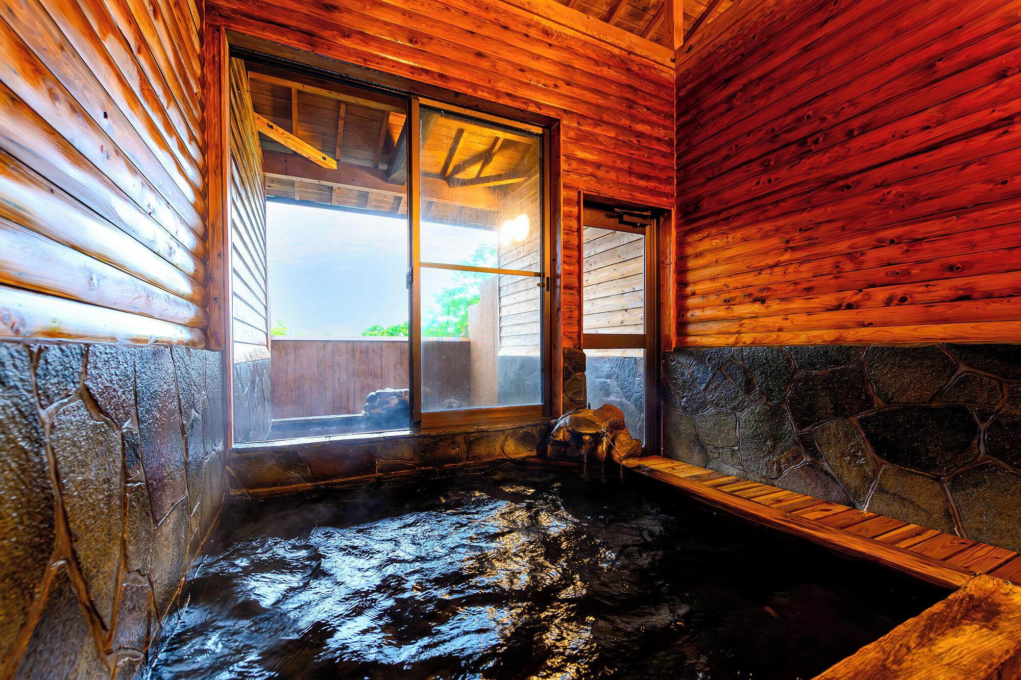 Interior de una cabaña de madera con un jacuzzi de paredes de piedra y un ventanal grande.