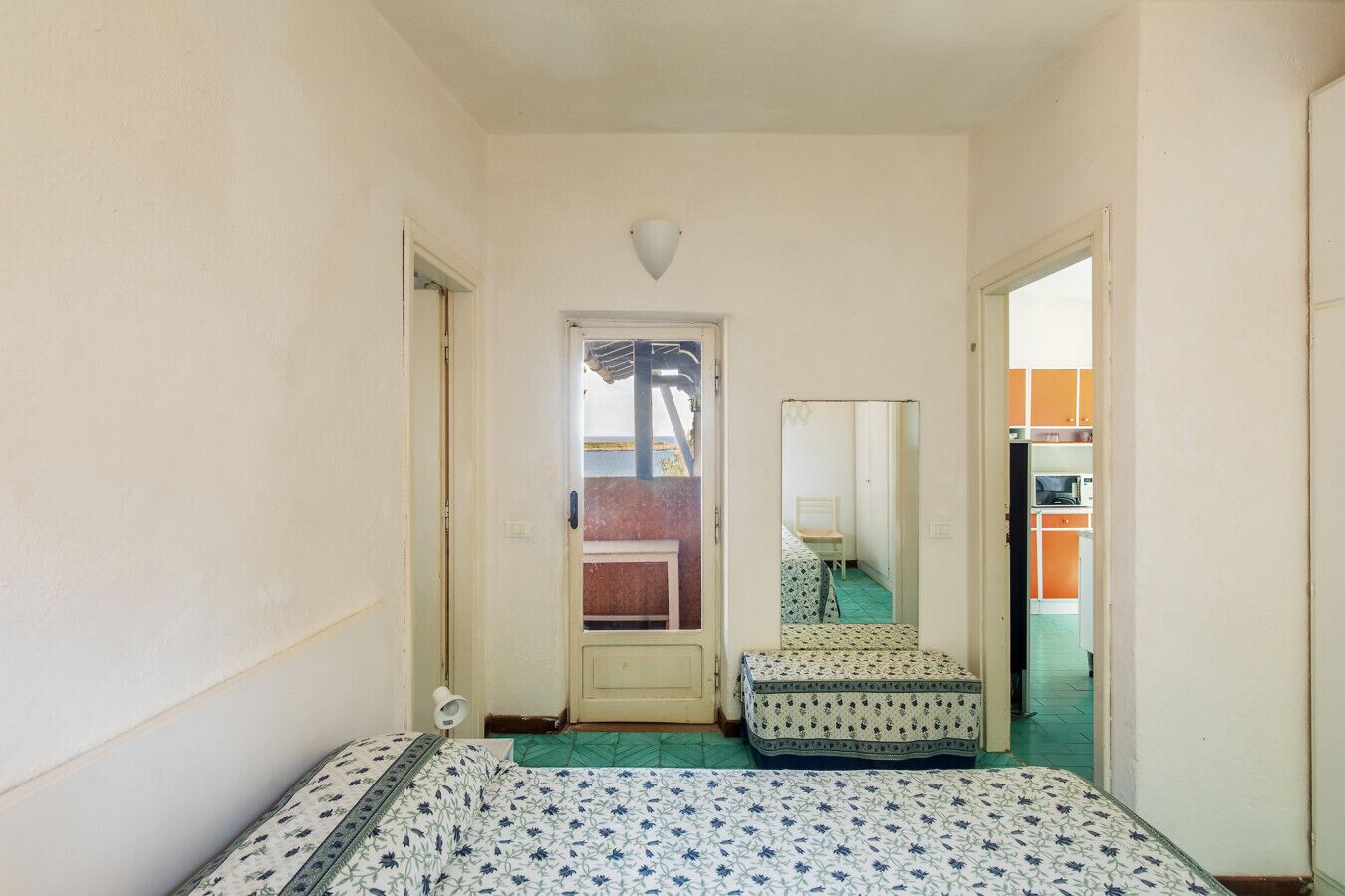 Habitació petita i estreta amb un llit, una taula petita i un mirall. Hi ha una porta que condueix a una altra habitació amb una cuineta.