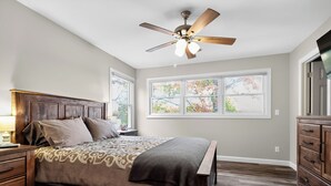 Une chambre à coucher comprenant un lit, une tête de lit en bois, des tables de chevet, une commode et un ventilateur de plafond.