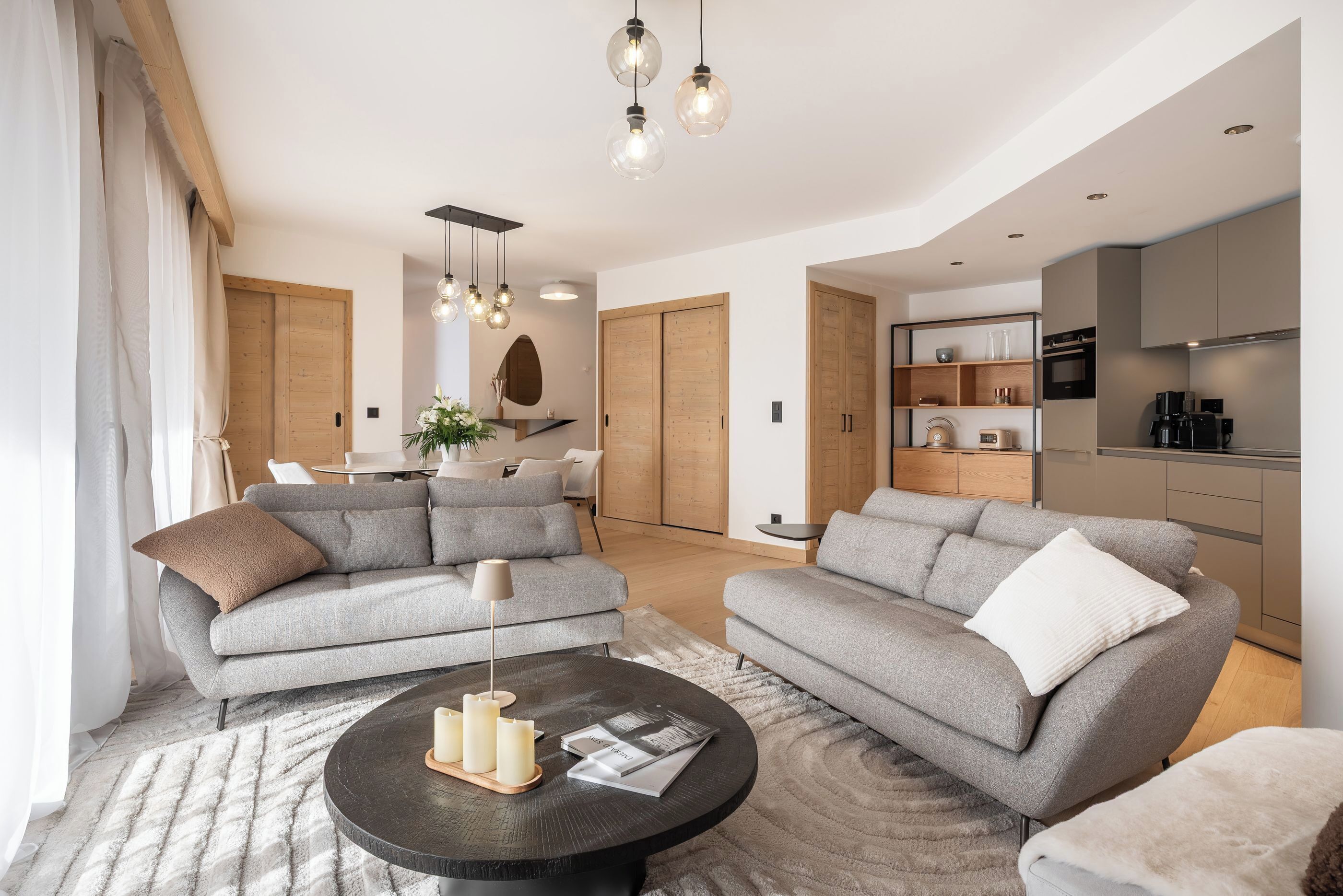 A modern living room with two grey sofas, a round coffee table with candles, and a dining area in the background.