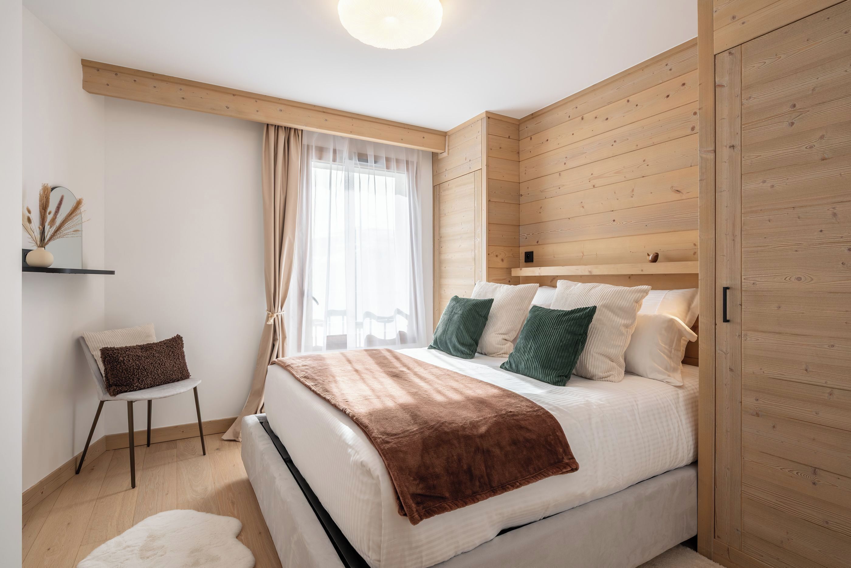 A bedroom with a bed, a chair, a wooden wardrobe, and a window with curtains.