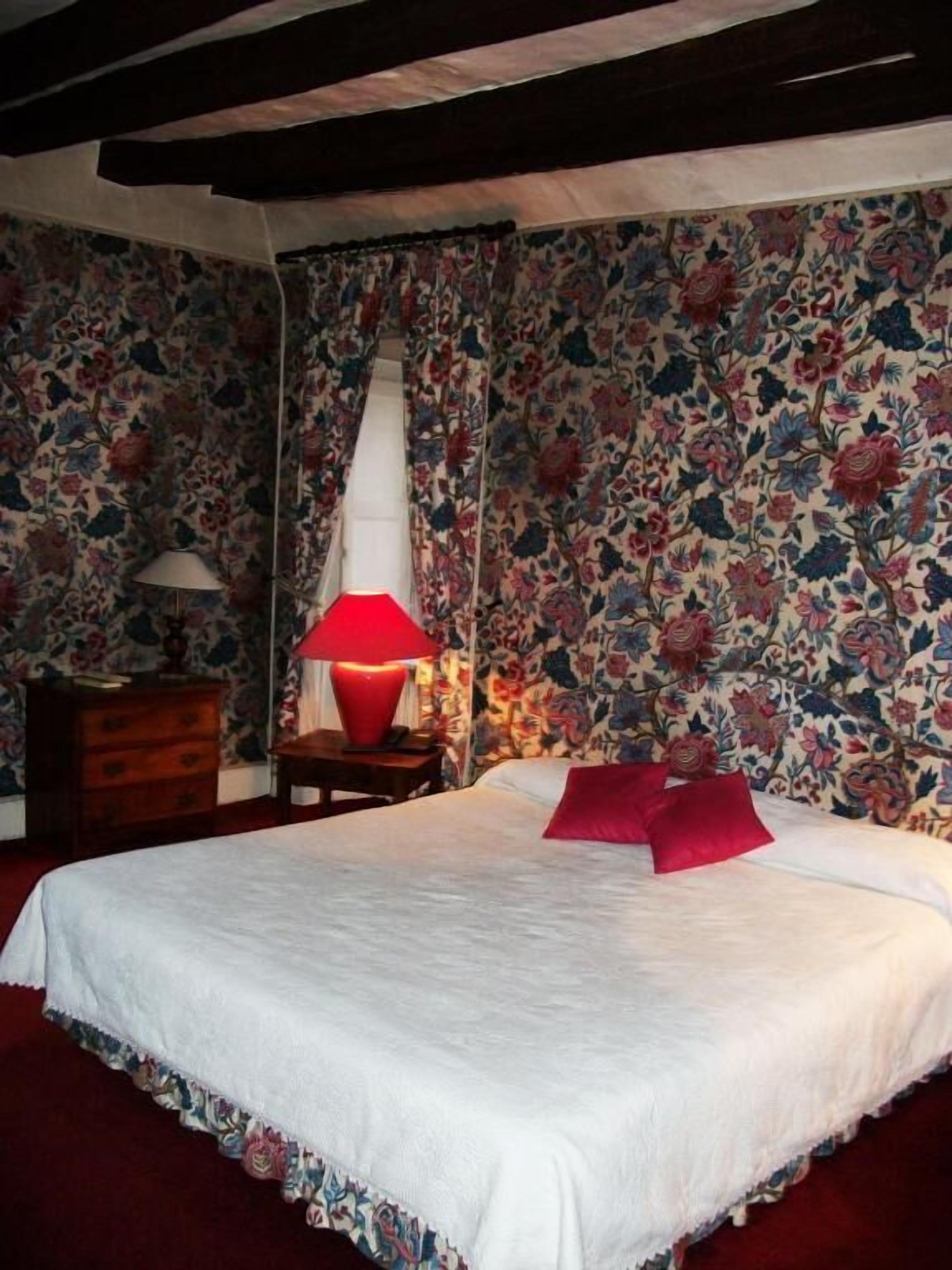 A bedroom with a floral wallpaper, a wooden dresser, a red lamp, and a bed with white bedding and red pillows.