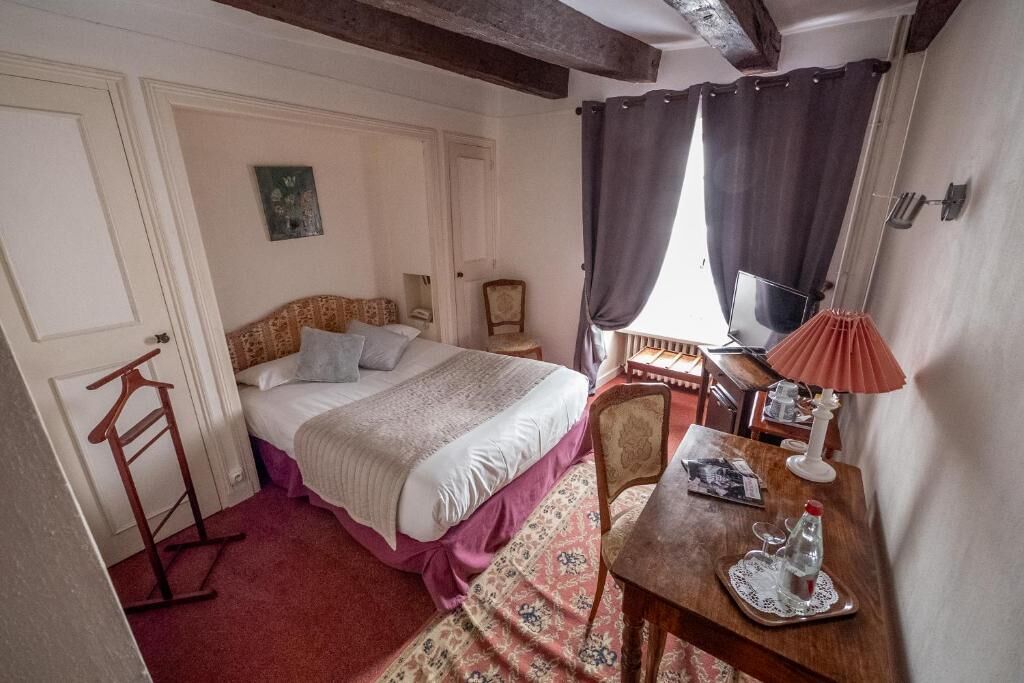 A bedroom with a bed, a wooden chair, a table with a lamp, and a window with curtains.