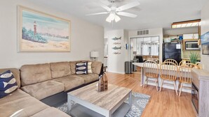 A modern living room with a sofa, coffee table, and dining area with chairs. There is a ceiling fan, a kitchen with a refrigerator, and a painting on the wall. - Walk to the Beach or the Bay! (Ocean City)