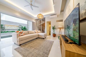 A modern living room with a sectional sofa, a large flat-screen TV, a wooden entertainment unit, a spiral staircase, and a large window. - New Modern, 2Br Villa in Canggu/Pool/Rooftop (Tabanan)