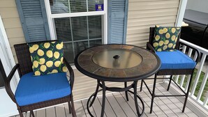 Une table ronde avec un plateau en verre, deux chaises en osier avec des coussins bleus, et une fenêtre avec des volets.
