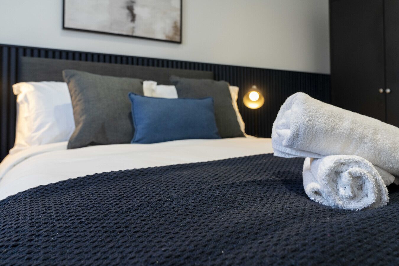 A neatly made bed with a dark textured blanket, a white pillow, and a rolled white towel.