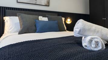 A neatly made bed with a dark textured blanket, a white pillow, and a rolled white towel.