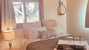Ein gemütliches Schlafzimmer mit Holzbalkendecke, einem Bett mit grüner Bettdecke, einem Nachttisch mit Lampe und einem Fenster mit Blick auf Bäume.