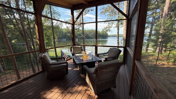 Een overdekt terras met rieten meubels, een tafel met glazen blad, en uitzicht op een meer omringd door bomen.
