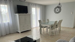 A modern living room with a glass coffee table, a white sofa, a dining table with chairs, a TV on a cabinet, and a wall clock. - Gîte des 3 Moulins - A haven of tranquillity in the heart of the vineyards (Gornac)