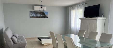 Una sala de estar moderna con una mesa de comedor de vidrio, sillas blancas, un sofá gris, un televisor montado y un cuadro en la pared.