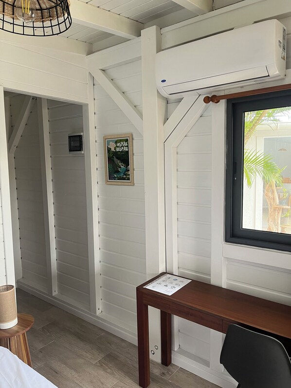 A room with white paneled walls, a wooden table, a chair, a window with a view of palm trees, and an air conditioning unit. - Coco Banana - Superior bungalow - pool view (Baie-Mahault)