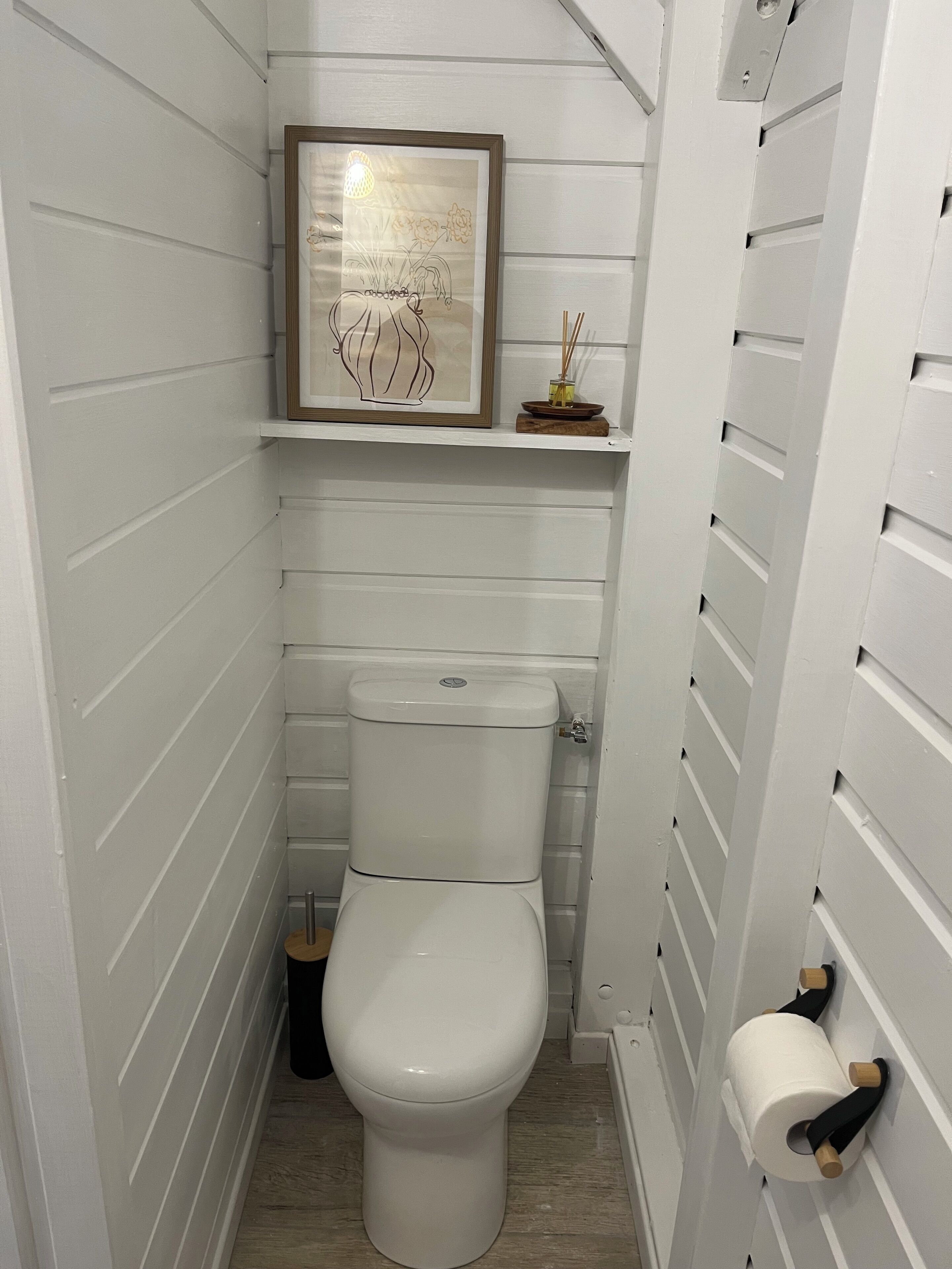 Un bagno piccolo con pareti bianche, dotato di un water, un'opera d'arte incorniciata sopra il water e uno scaffale con un piccolo oggetto decorativo.