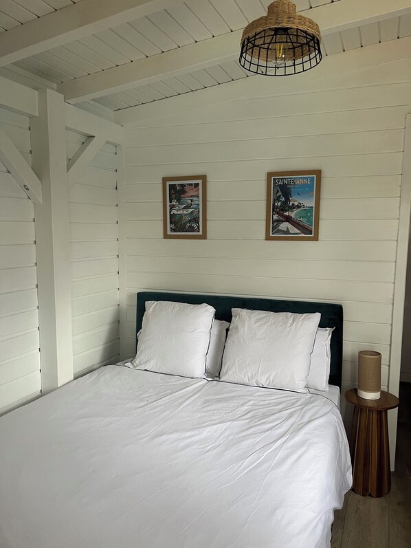 A bedroom with a bed, white bedding, a wooden headboard, two framed pictures, a hanging light fixture, and a bedside table with a lamp. - Coco Banana - Superior bungalow - pool view (Baie-Mahault)