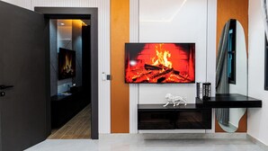A modern living room with a large flat-screen TV mounted on a white wall, a black entertainment unit below, and a mirror on the right.