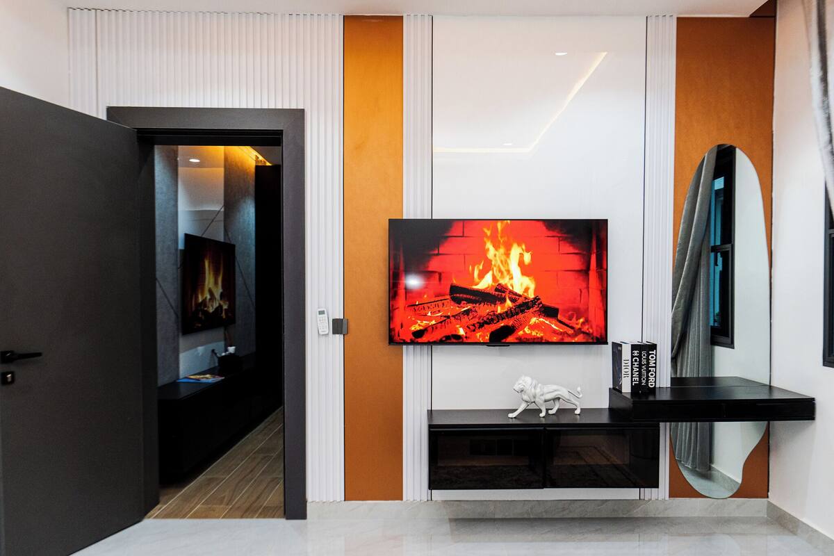 A modern living room with a large flat-screen TV mounted on a white wall, a black entertainment unit below, and a mirror on the right.