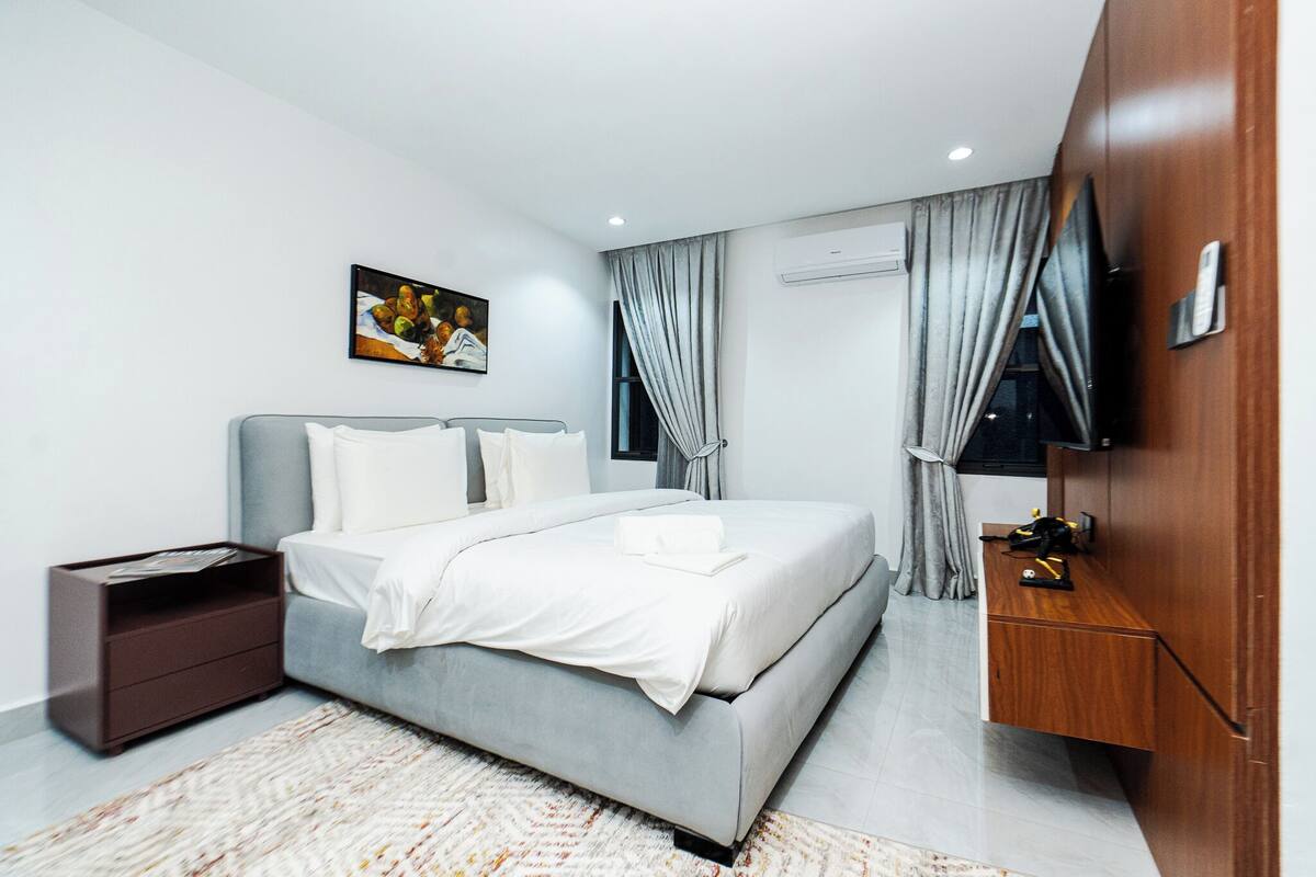 A modern hotel room with a grey bed, white linens, a wooden nightstand, a TV on a wooden cabinet, and a painting on the wall.