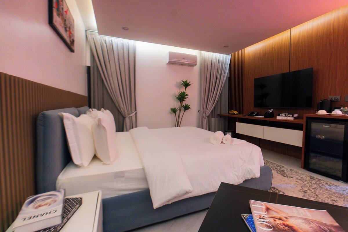 A modern hotel room with a bed, a TV, a minibar, and a desk.