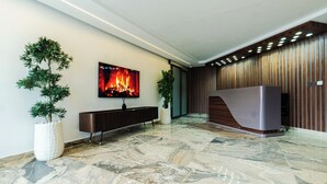 A modern lobby with a marble floor, a large flat-screen TV, a dark wooden reception desk, and two potted plants.