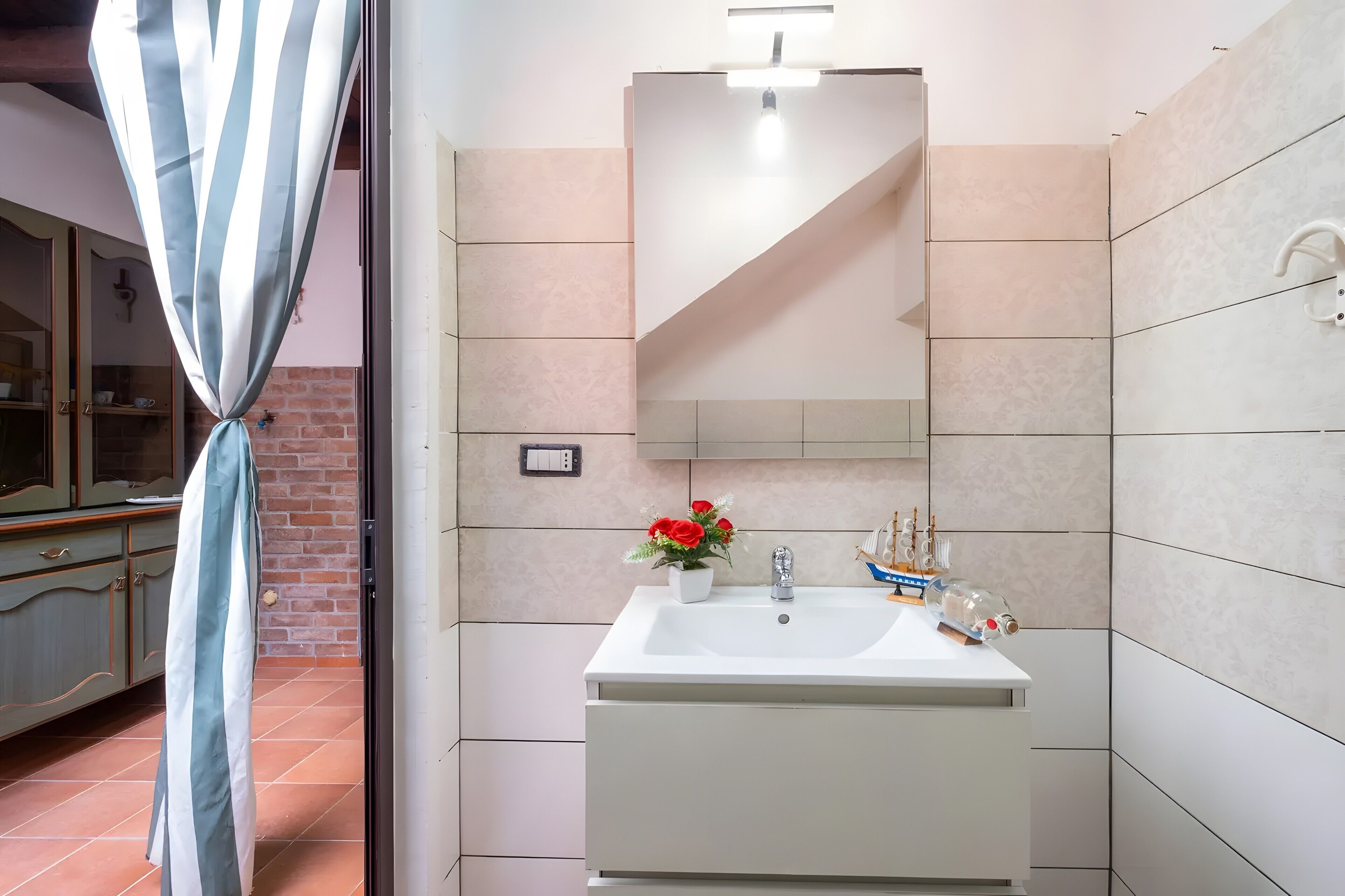 Un baño con un lavamanos blanco, un espejo y una planta en maceta con flores rojas.