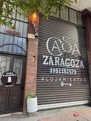 A closed storefront with a large metal shutter, a sign reading 'CASA ZARAGOZA ALOJAMIENTOS,' and a contact number '3951151579.' - Hotel Casa Zaragoza, San Juan de los Lagos (San Juan de los Lagos)