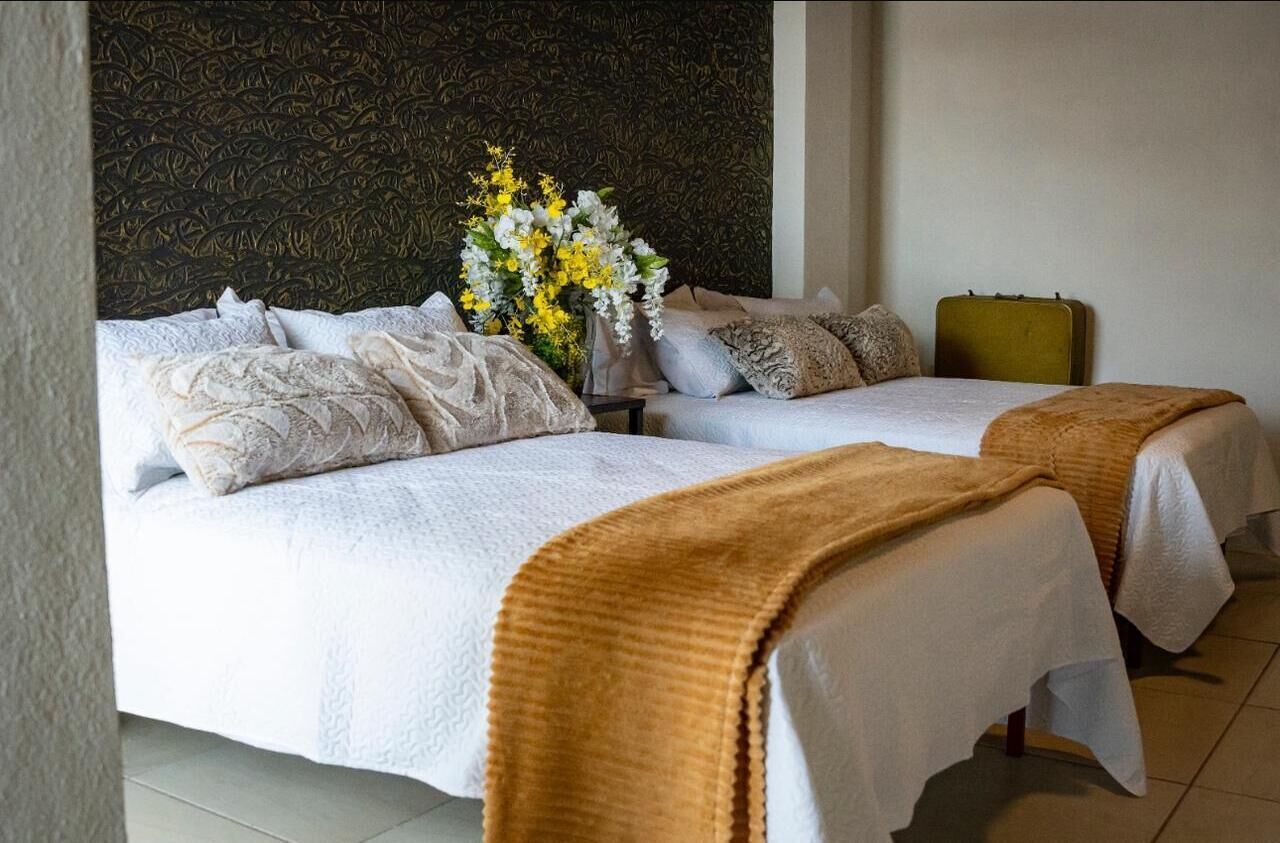 Ein Hotelzimmer mit zwei Betten, einem dunkel gemusterten Kopfteil, einer Vase mit Blumen und einem Koffer.