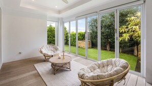 A spacious room with large sliding glass doors leading to a well-maintained garden. There are two wicker chairs and a small round table with a candle. - Modern Beach Retreat | Eden Beach Sanctuary (Jindalee)