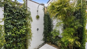 A small, enclosed shower area with a white tiled wall, a showerhead, and lush green plants growing on the walls. - Modern Beach Retreat | Eden Beach Sanctuary (Jindalee)