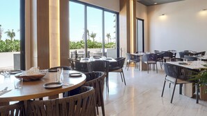 Un comedor amplio con mesas y sillas redondas, ventanas grandes y vistas a palmeras.