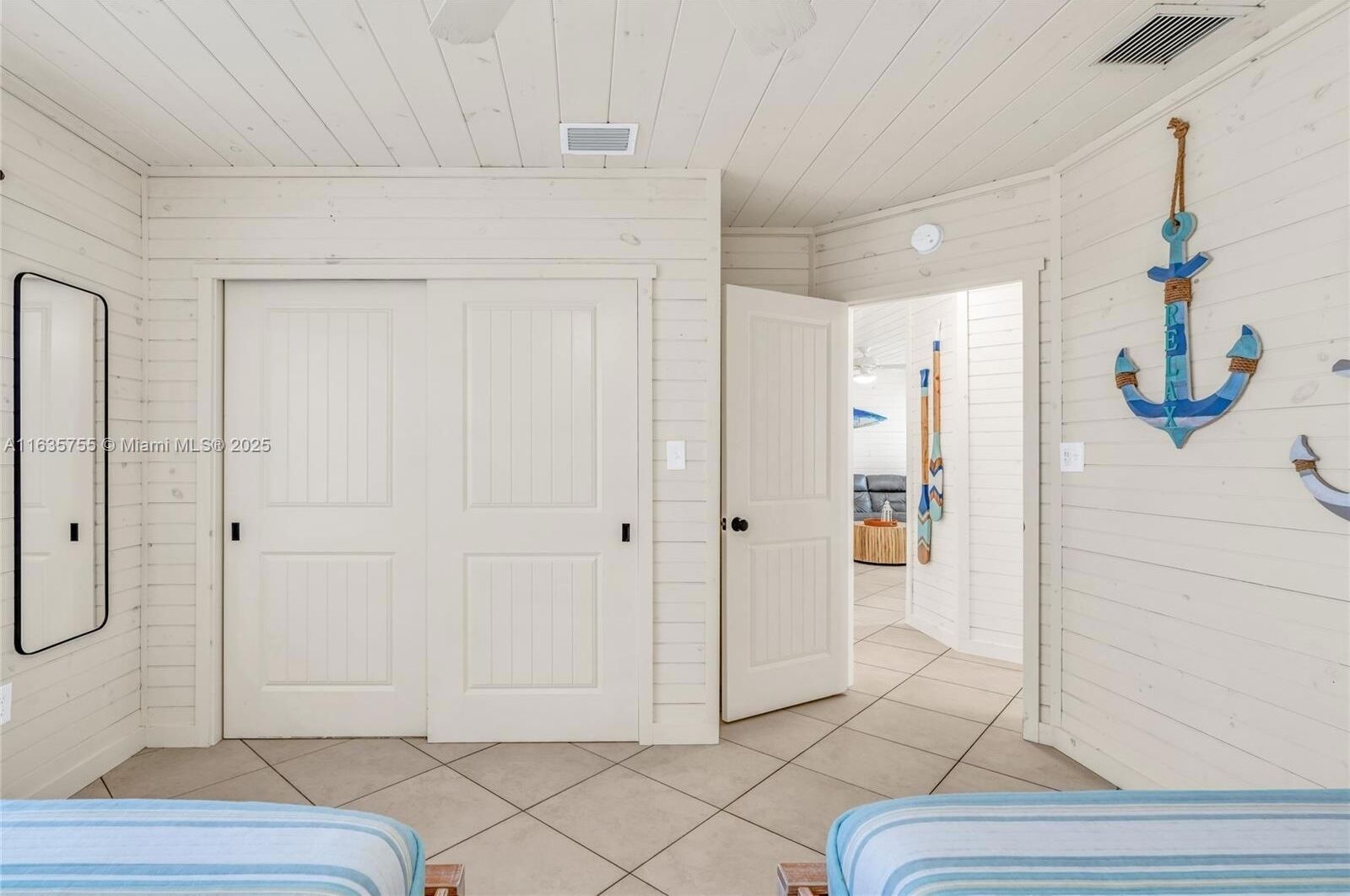 A room with two beds, white wooden walls, and a hallway leading to another room.