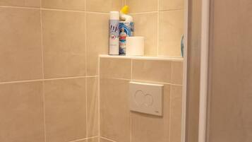 A small, enclosed shower stall with a toilet, a shelf with toiletries, and a control panel.
