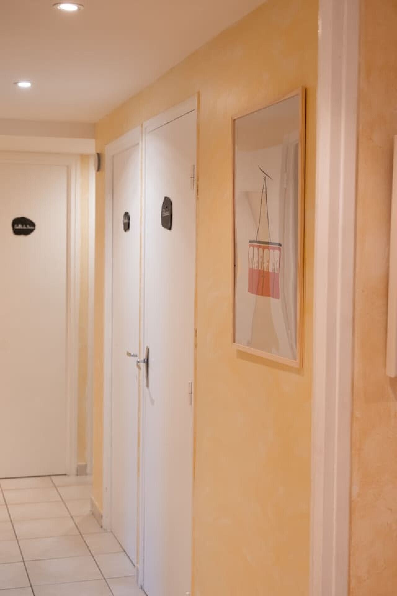Un couloir avec deux portes blanches, dont l’une porte une enseigne noire, et un tableau encadré accroché au mur.