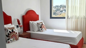 Een hotelkamer met twee bedden, elk met een rode hoofdbord en witte beddengoed. Er is een groot raam met uitzicht op een stadsgezicht.