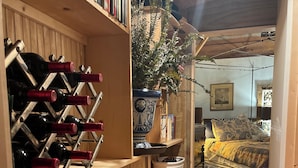 A cozy room with a bookshelf, a wine rack, a large vase with a plant, a patterned rug, and a bed with a patterned bedspread.