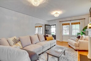 Ein geräumiges Wohnzimmer mit einer großen Couch, einem Couchtisch und einem Kamin. Zwei Fenster mit Vorhängen und eine Pendelleuchte sind ebenfalls vorhanden.