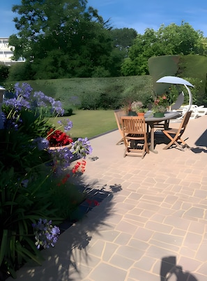 Une terrasse extérieure avec une table et des chaises, entourée d’un jardin où poussent différentes plantes et fleurs.