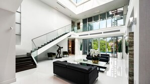 A spacious, modern interior with a high ceiling, a staircase, a living area with a sofa and coffee table, and large windows offering a view of the outdoors. - Luxury 7BR Waterfront Villa w/ Rooftop, Pool & Dock – Sleeps 14 near Las Olas (Fort Lauderdale)