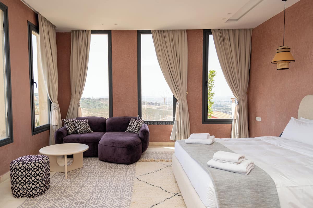 Una habitación de hotel moderna con una cama grande, un sofá, una mesita redonda y un otoman con estampado.